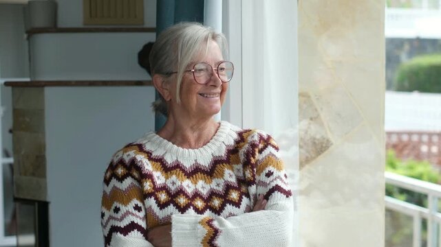 Smiling mature woman wearing eyeglasses and knitted sweater, standing indoors with crossed arms while peacefully looking out the window, enjoying retirement in a bright cozy house