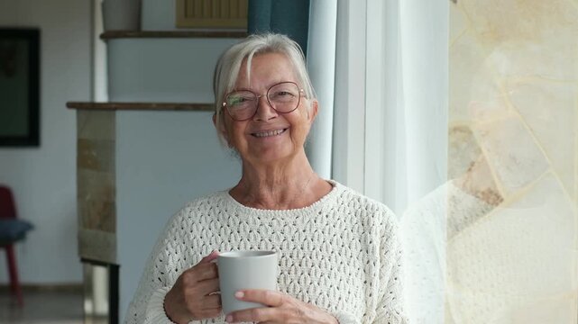 Elderly woman smiling warmly while holding coffee cup near window curtain, enjoying peaceful morning beverage indoors in her comfortable home setting