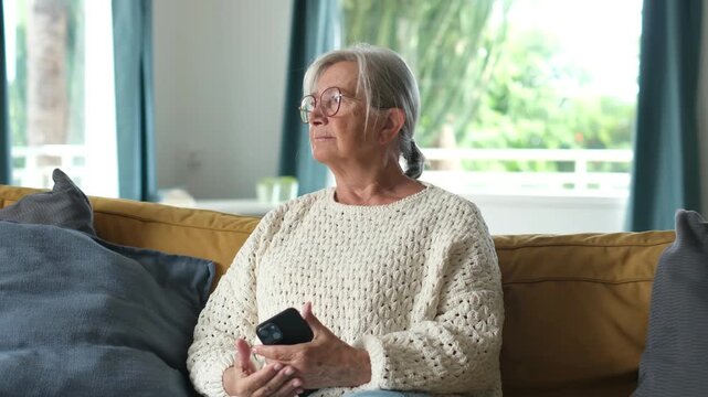 Elderly woman with white hair and eyeglasses sitting comfortably on a living room couch, holding a smartphone and gazing thoughtfully out the window in a calm, reflective moment at home