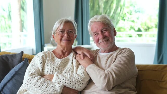 Happy senior couple sitting closely on a cozy sofa at home, looking at the camera with warm smiles and enjoying peaceful retirement together in their bright living room