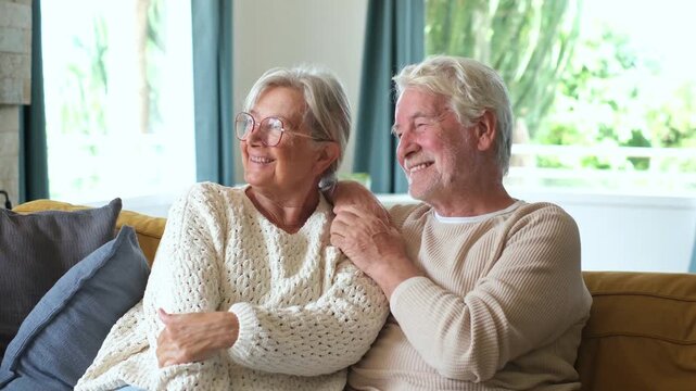 Smiling elderly husband and wife sitting together on comfortable couch in living room, talking cheerfully and looking out the window while enjoying quiet retired lifestyle