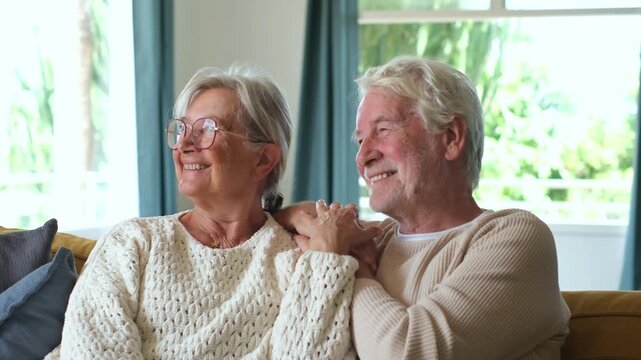 Loving senior couple smiling, embracing, and daydreaming while sitting together on a comfortable sofa in their bright modern living room, enjoying retirement life