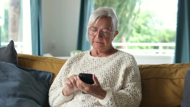 Focused elderly grandmother wearing glasses relaxing on a cozy couch at home while typing messages and browsing the internet on her mobile phone, enjoying modern lifestyle indoors