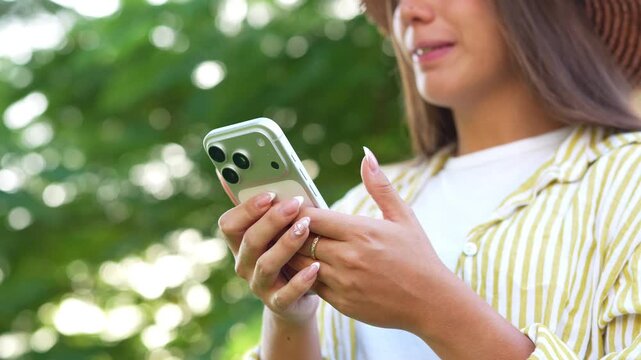 Young smiling woman wearing straw hat using smartphone while standing outdoors in city park, browsing internet, texting messages, and enjoying sunny summer day