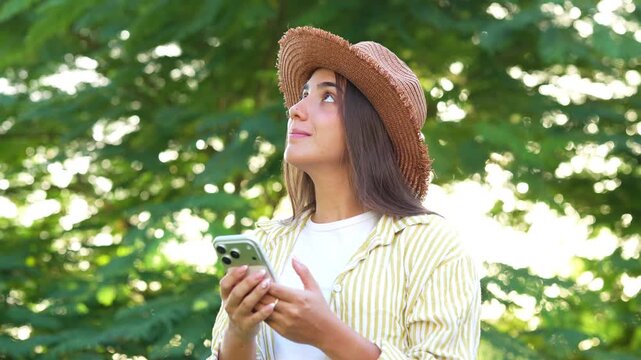 Young female traveler wearing stylish straw hat using modern smartphone, texting online and admiring nature while standing in green city park on bright summer day