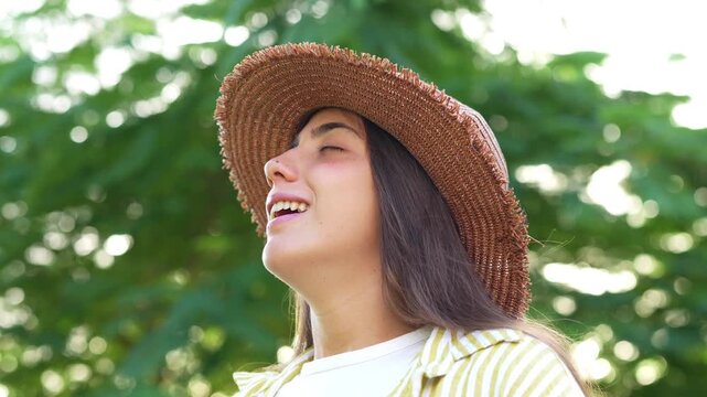 Young woman in a straw hat, smiling with eyes closed and taking a deep breath of fresh air while enjoying nature in a sunny summer park