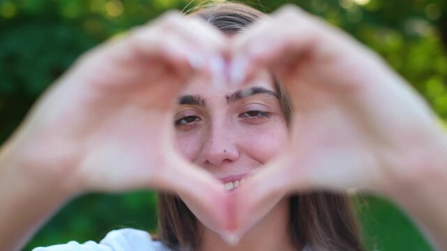 Happy young brunette woman smiling and making a heart shape sign with her hands outdoors in a sunny green park, expressing love, affection, positivity and gratitude to camera