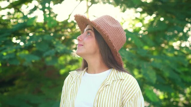 Young woman in a straw hat, smiling at the camera and looking around in a sunlit green park, enjoying a cheerful, relaxed summer day outdoors among trees and foliage