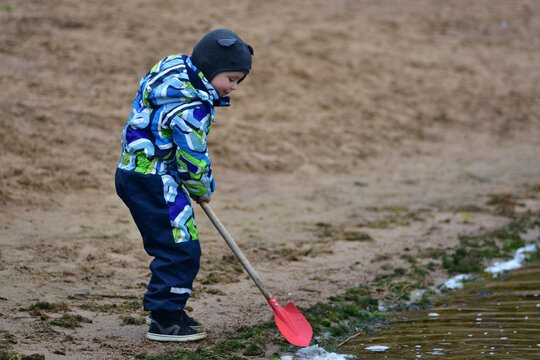 Young boy wearing warm outdoor clothing using a red shovel at the edge of a sandy lakeside beach, exploring water, sand and nature during a cool autumn outdoor activity.