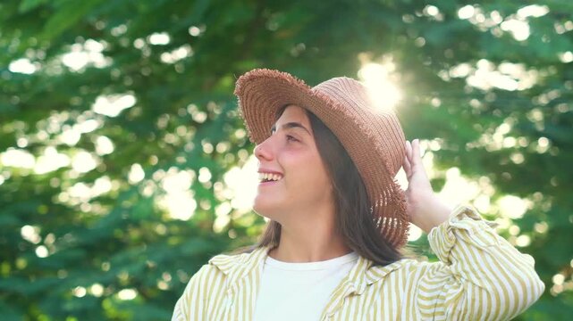 Cheerful young woman in a straw hat looking around and smiling in a lush green park, enjoying warm summer sunlight filtering through leafy trees