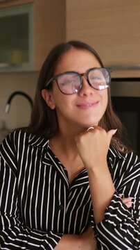Confident young businesswoman wearing eyeglasses, smiling at the camera and resting her chin on hand while daydreaming in modern home kitchen setting