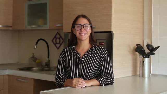 Smiling young woman in eyeglasses leaning on a modern kitchen counter, daydreaming and looking around with satisfaction in a cozy, sunlit home интерьер