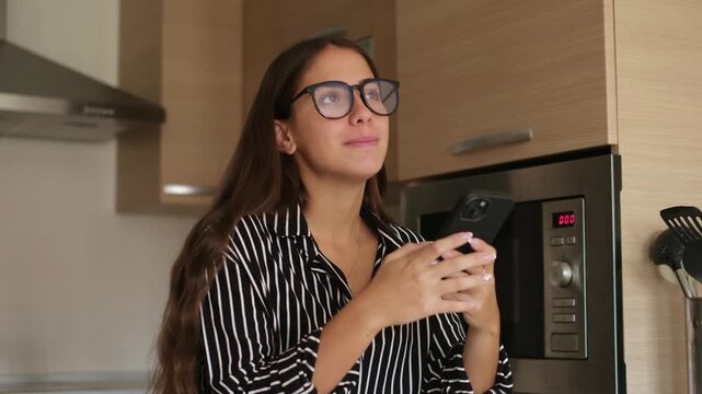 Cheerful young brunette woman wearing eyeglasses standing in modern domestic kitchen while texting on smartphone, browsing social media and smiling thoughtfully