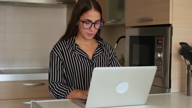 Focused young businesswoman wearing glasses typing on laptop while working remotely in modern kitchen, daydreaming and looking up with a gentle smile