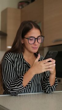 Young woman wearing glasses and smiling while standing in a modern home kitchen, holding a smartphone and texting friends or browsing social media