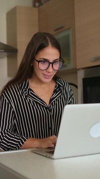 Cheerful young woman with eyeglasses typing on a laptop computer while standing at the kitchen counter, smiling and working remotely from home during daytime