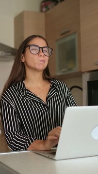 Thoughtful female freelancer wearing eyeglasses typing on notebook keyboard, pausing to think and touching chin while working remotely in modern home kitchen