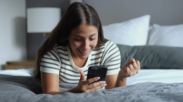 Young hispanic woman lying on a cozy bed, smiling with surprise and joy while reading exciting online news on her smartphone in a relaxed bedroom setting