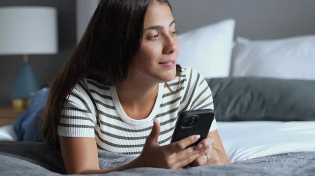 Young relaxed woman lying down on comfortable bed in cozy bedroom, smiling while typing text message on smartphone and looking away thoughtfully, enjoying evening leisure time at home