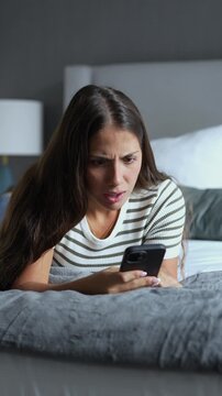 Annoyed young woman lying on a comfortable bed at home, reading unpleasant messages on her smartphone and gesturing with frustration while dealing with bad digital news