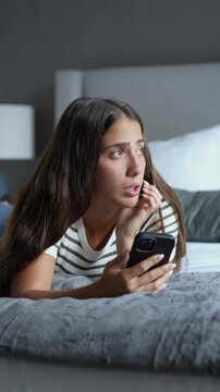 Melancholic young woman lying on bed at night, holding smartphone while looking sad, disappointed and checking social media or waiting for message indoors