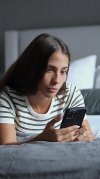 Young brunette woman lying on her bed in a dim bedroom, browsing and typing on her smartphone with a concentrated expression at night