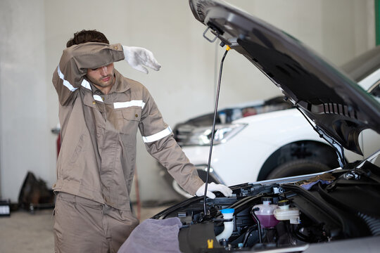 A tired mechanic wiping sweat from his forehead while working on a car engine in a garage.