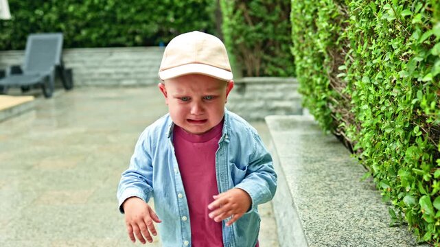 A toddler wearing a white cap, blue shirt, and pink clothes stands on a stone path beside a green hedge in an outdoor garden.