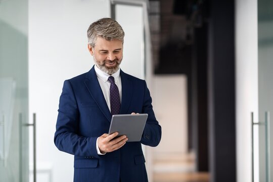 Smiling Caucasian Man In Suit Engaged With Tablet While Standing In Modern Office Environment, Reflecting Focus And Professionalism In A Corporate Setting, Business Concept.