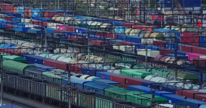 Freight wagons tank cars and shipping containers form parallel rows beneath a grid of overhead railway wires with weathered open wagons and green covered cars prominent in the elevated close view