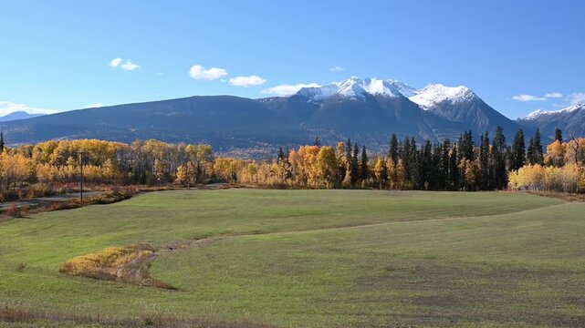 The view pans right across a green field bordered by golden deciduous trees and dark conifers, with snowcapped mountains rising beneath a blue sky.