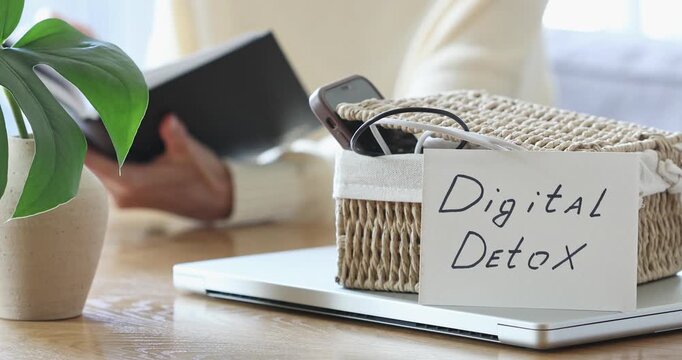 Digital detox concept from woman reading paper book while phone and laptop gadgets lying offline. Home and mental leisure instead social media and technology addiction.