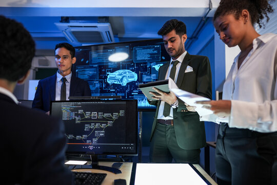Cinematic view of automotive software engineers comparing printed specs in front of the world map analytics beside a screen glowing with a car wireframe blueprint