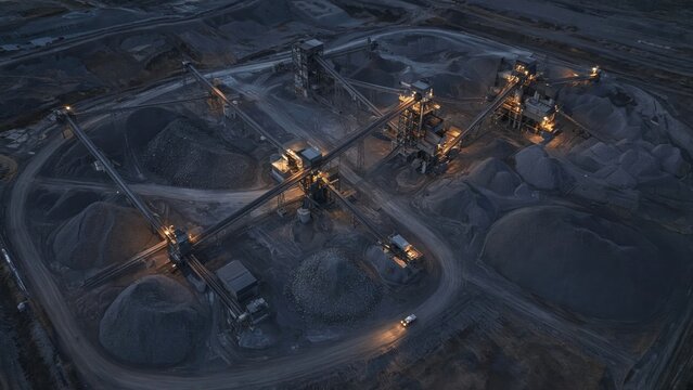 Sprawling mineral processing site at blue hour with glowing conveyors and dark stockpiles