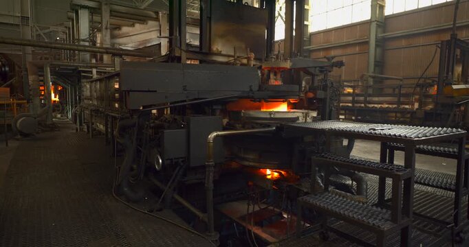 Glass container factory feeding station shines orange amid pipework and heavy equipment with grated access steps in the foreground and high windows above the workshop in a wide stationary view