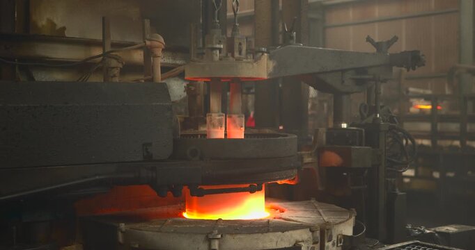 Glass feeding machinery stands over an incandescent furnace outlet with two vertical rods and a horizontal arm framed by dark pipes and a factory interior as the view lowers toward the opening