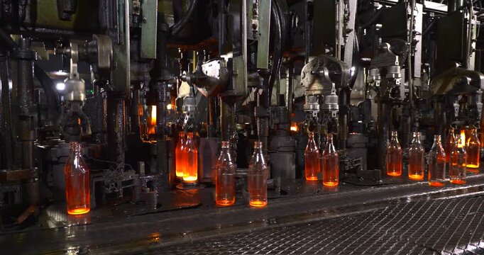 Hot glass containers are transferred at forming stations with glowing bases lined across the foreground and rows of moving neck grips receding beside a grated walkway in the bottle factory