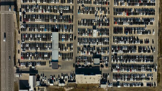 Top-down aerial view of a large vehicle storage yard containing hundreds of cars arranged in organized rows. The paved parking area features access lanes, vehicle inventory