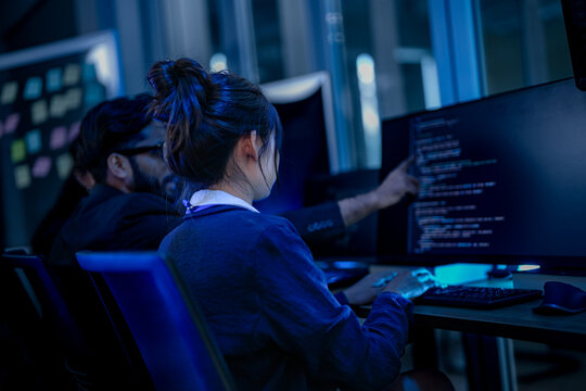 Software developers pointing at a desktop monitor to analyze artificial intelligence source code and debug deep learning software during a late night tech operation.