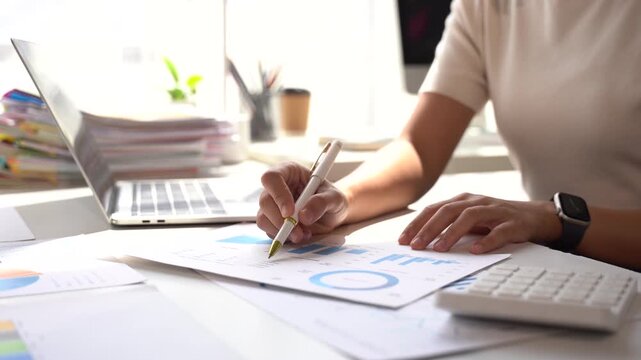 Financial Analysis at Desk: A focused individual meticulously analyzes financial reports and charts on a desk, illuminated by natural light streaming through a window.