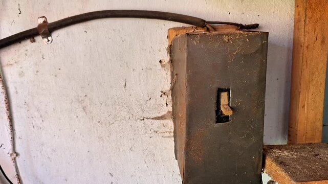 An old metal electrical junction box mounted on a cracked, weathered wall with exposed cables and worn wiring. The scene highlights deteriorating electrical infrastructure in an aging building