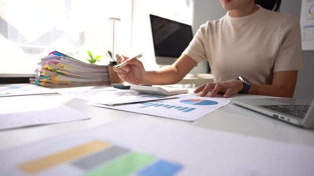Businesswoman Calculating Financial Data: A focused businesswoman meticulously analyzes financial reports, using a calculator at her desk, surrounded by stacks of documents and a computer.