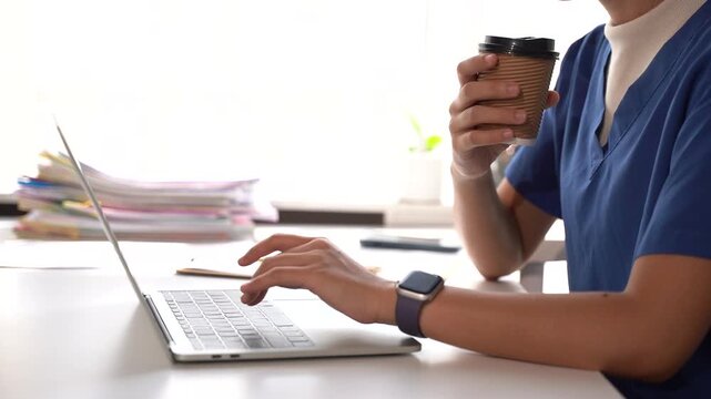 Doctor Working and Drinking Coffee: A medical professional in blue scrubs types on a laptop, holding a paper cup of coffee, symbolizing a busy day in healthcare.