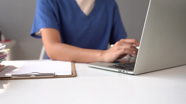 Doctor Typing on Laptop: A medical professional wearing blue scrubs is focused on typing on a laptop computer, with a clipboard and medical chart on the desk in front of them.