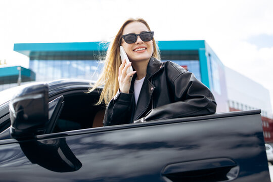 Young woman in sunglasses leaning out of a car window, smiling and using her smartphone for a conversation in the city