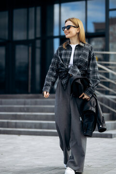 Young woman smiling, walking on city street, wearing plaid shirt, wide-leg trousers, sunglasses, carrying a jacket