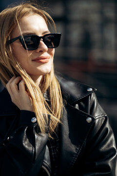 Young woman smiling while looking away, wearing a stylish black leather jacket and sunglasses in urban environment