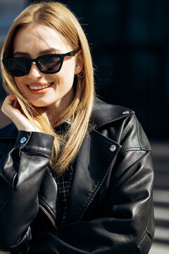 Blonde woman wearing sunglasses and a black leather jacket, smiling and enjoying urban fashion in natural sunlight