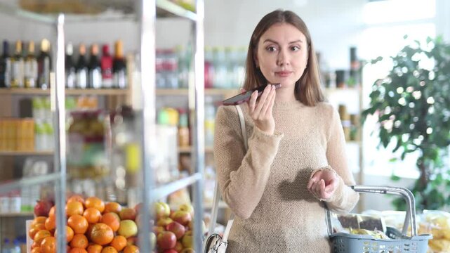 Portrait of young girl with shopping basket choosing products in supermarket while recording audio message on mobile phone 