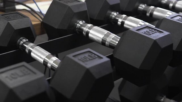 A close sliding view passes black rubber hex dumbbells with textured metal handles on a rack.